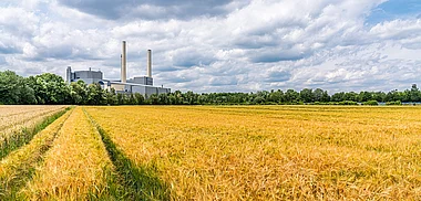Industrieanlagen mit Schornsteinen dominieren eine grüne Landschaft mit Getreidefeldern.